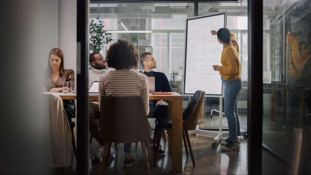 A woman in business casual stands in front of a digital whiteboard pointing out a piece of information in what appears to be a workflow. A table of colleagues are giving her their attention.