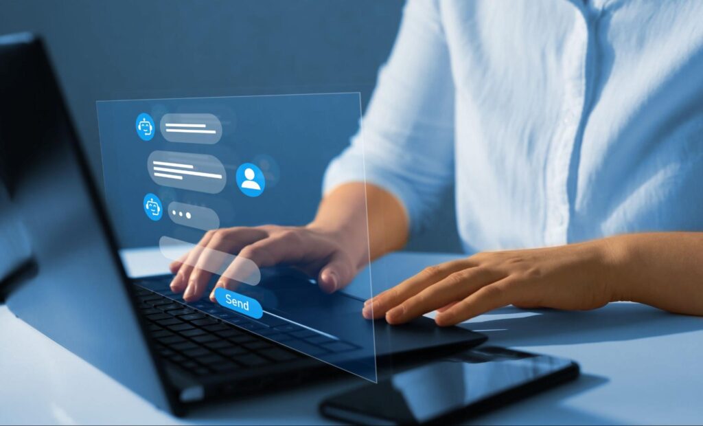 A person wearing a light blue button down sits at their laptop, their hands lightly touch the keys. A digital overlay appears to come from the screen showing that our person is chatting with an AI chatbot. 