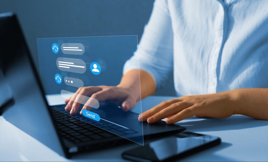 A person wearing a light blue button down sits at their laptop, their hands lightly touch the keys. A digital overlay appears to come from the screen showing that our person is chatting with an AI chatbot.